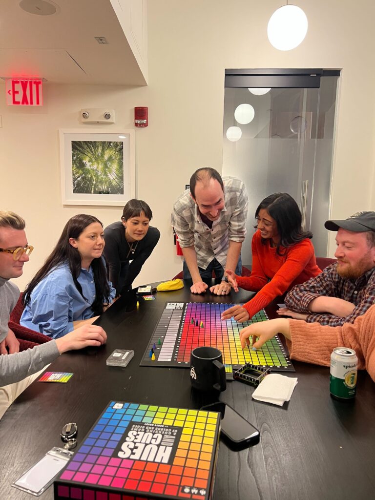 Six team members gathered around a table playing the game Hues and Cues