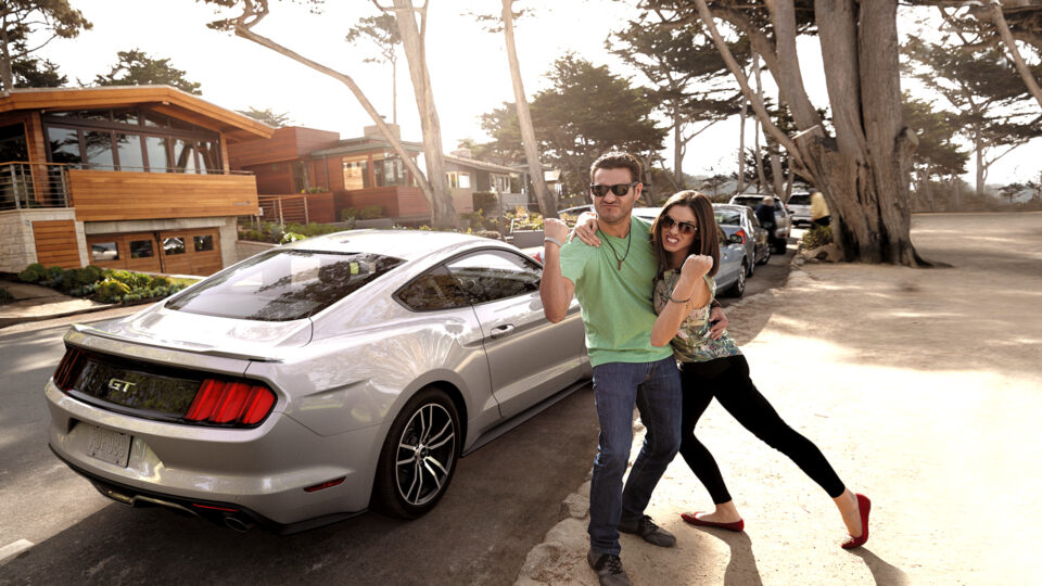 Two young people strike a pose with fists in the air outside a Ford Mustang on a tree lined street