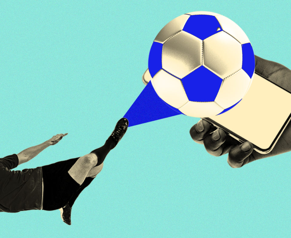 Soccer player kicking a soccer ball into the direction of a hand holding a cellphone.